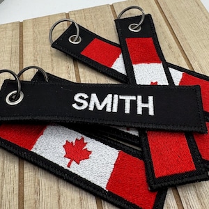 Peut inclure: Un ensemble de porte-clés noirs et rouges avec un motif de drapeau canadien, l'un des porte-clés portant le nom "SMITH" brodé.