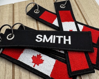 May include: A set of rectangular fabric keychains featuring the Canadian flag design in red and white, with the name SMITH embroidered in white thread on a black background. Each keychain includes a silver metal grommet and a split ring.