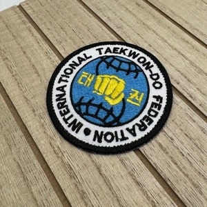 Può includere: Una toppa ricamata bianca, nera e blu con il testo "International Taekwondo Federation" e un pugno all'interno di un globo.