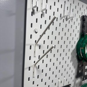 May include: White pegboard with various white hooks and accessories. The pegboard has evenly spaced holes for attaching the hooks. Some hooks are angled, and others are straight. The background is a light gray wall.