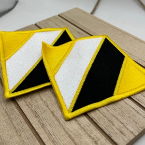 May include: Two yellow and black embroidered patches in the shape of a diamond. The patches have a white diagonal stripe.