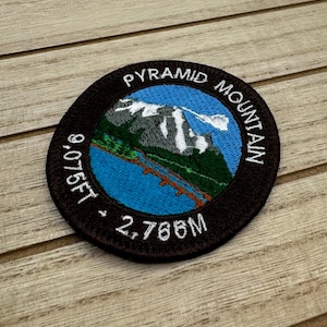 May include: Black and white embroidered patch with a blue and green mountain scene. The text "PYRAMID MOUNTAIN 9,075FT - 2,766M" is embroidered around the mountain scene.