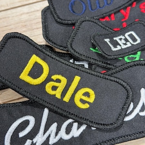 May include: A collection of black embroidered name patches. Names are stitched in various colours, including yellow, blue, and white. Names visible are "Dale", "Olivia", "Leo", and "Charles". The patches are displayed on a wooden surface.