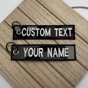 Puede incluir: Dos llaveros negros y de camuflaje con anillas plateadas. Un llavero dice "CUSTOM TEXT" en letras blancas, y el otro dice "YOUR NAME". Los llaveros son rectangulares.
