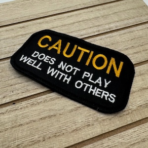 May include: Black embroidered patch with white text that reads "CAUTION DOES NOT PLAY WELL WITH OTHERS". The word "CAUTION" is in yellow.