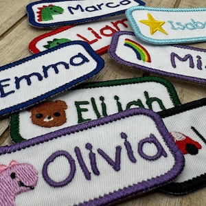 May include: A collection of colourful embroidered name tags with various designs, including a dinosaur, a rainbow, a bear, and a car. The names on the tags are Emma, Liam, Marca, Elijah, Olivia, Mica, and Isobel.