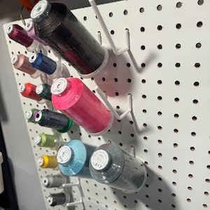 May include: A white pegboard with various spools of thread in different colors, including black, pink, teal, and gray. The spools are labeled with numbers, such as "817" and "2340".