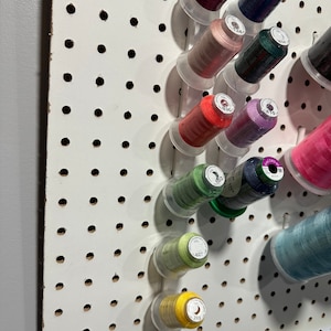 Puede incluir: Un tablero de clavijas blanco con varios carretes de hilo de diferentes colores, incluyendo rojo, rosa, verde, azul y plateado. Los carretes están organizados en filas y están etiquetados con números.