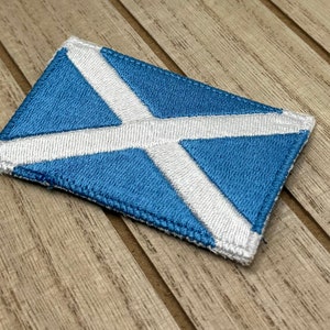 Puede incluir: Un parche bordado con la bandera de Escocia, el Saltire, con una cruz blanca sobre fondo azul.