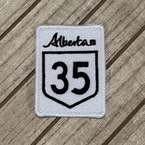 Peut inclure: Un écusson rectangulaire blanc avec le mot "Alberta" en écriture noire au-dessus d'un bord noir en forme de bouclier contenant le numéro "35" en gras noir. L'écusson a une bordure cousue.