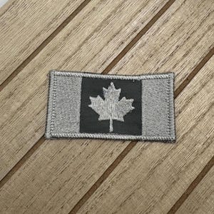 Op de afbeelding: Een klein, geborduurd patch met de Canadese vlag. De vlag is een rood esdoornblad op een witte achtergrond met een zwarte rand. Het patch is gemaakt van grijs stof.