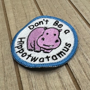Può includere: Una toppa ricamata bianca e blu con un ippopotamo rosa e il testo "Don't Be a Hippopotamus".