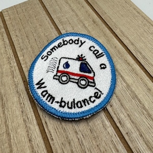 Può includere: Una toppa ricamata bianca con un bordo blu. La toppa presenta un'ambulanza di cartoni animati con il testo "Somebody call a wambulance".