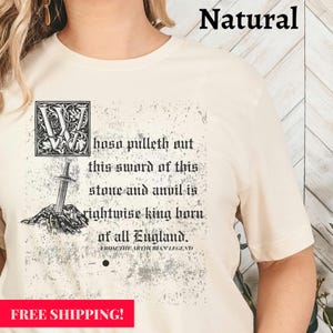 May include: A cream-coloured t-shirt featuring a medieval-style graphic. The design includes a sword in a stone, ornate letter, and text that reads "hoso pulleth out this sword of this stone and anvil is rightwise king born of all England. From the Arthurian Legend."