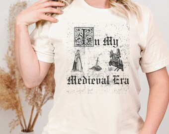 May include: A white t-shirt with a distressed graphic print of a medieval scene. The graphic includes a sword, a knight on horseback, a woman in a gown, and the text "I'm in my Medieval Era".