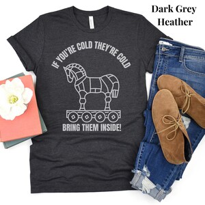 A dark grey heather shirt with the words: If your cold they're cold bring them inside! in a white simple font is lying on a white surface with jeans, boots & books. The words surround a line art image of the Trojan Horse.