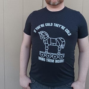 Man wearing a dark grey heather shirt with the words: If your cold they're cold bring them inside! in a white simple font. The words surround a line art image of the Trojan Horse.