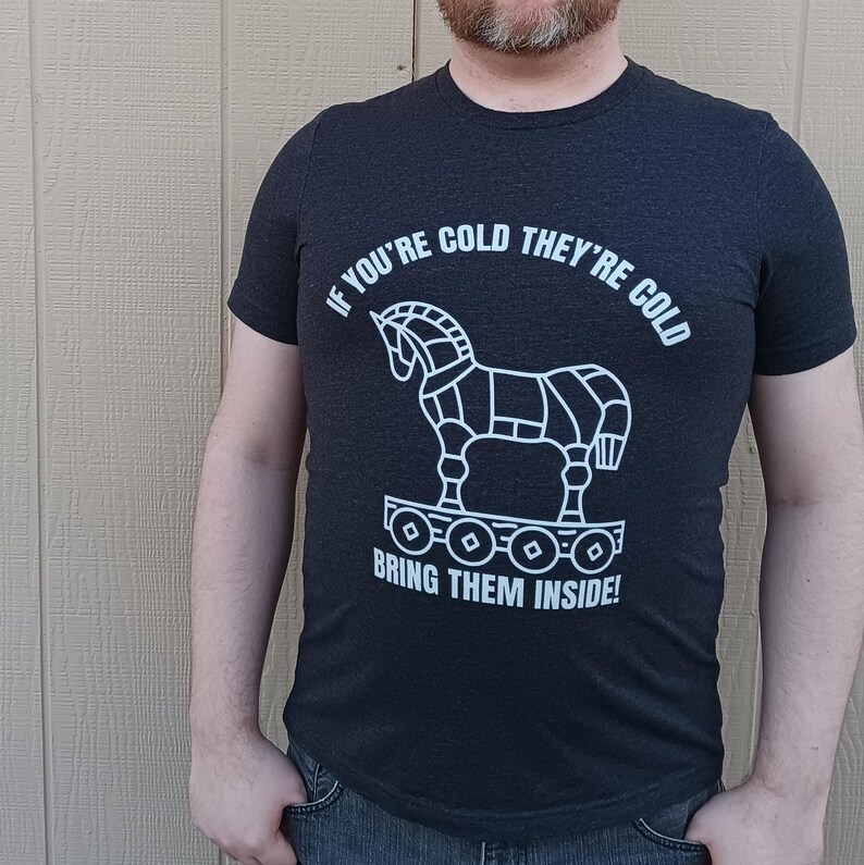 Man wearing a dark grey heather shirt with the words: If your cold they're cold bring them inside! in a white simple font. The words surround a line art image of the Trojan Horse.