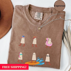 May include: Espresso-coloured t-shirt with a crew neck and short sleeves. The shirt features a graphic of six whimsical figures, each with a unique hat or accessory. The text "FREE SHIPPING!" is at the bottom.