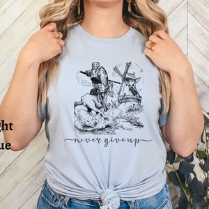 Woman standing against a wall wearing a light blue shirt with the words never give up in a simple cursive font of black. Above that is an illustration of Don Quixote fighting a windmill.