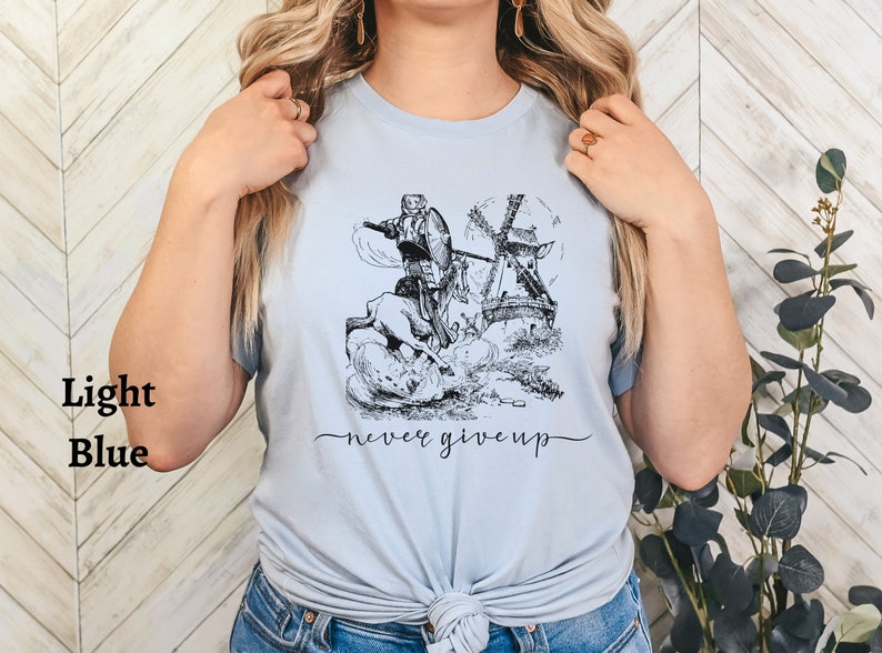 Woman standing against a wall wearing a light blue shirt with the words never give up in a simple cursive font of black. Above that is an illustration of Don Quixote fighting a windmill.