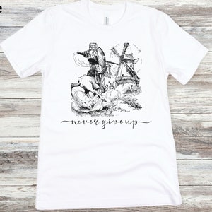 A white shirt with the words never give up in a simple cursive font of black is lying on a beige floor. Above that is an illustration of Don Quixote fighting a windmill.