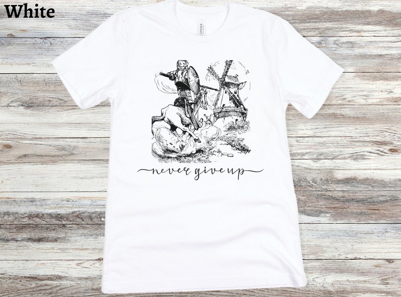 A white shirt with the words never give up in a simple cursive font of black is lying on a beige floor. Above that is an illustration of Don Quixote fighting a windmill.