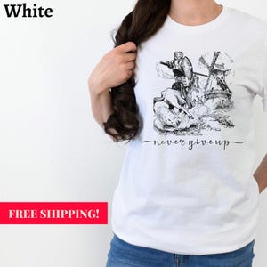 Woman standing against a wall wearing a white shirt with the words never give up in a simple cursive font of black. Above that is an illustration of Don Quixote fighting a windmill. Free shipping.