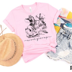 A pink shirt with the words never give up in a simple cursive font of black is lying on a white floor with shorts, straw hat, sunglasses & a scarf. Above that is an illustration of Don Quixote fighting a windmill.