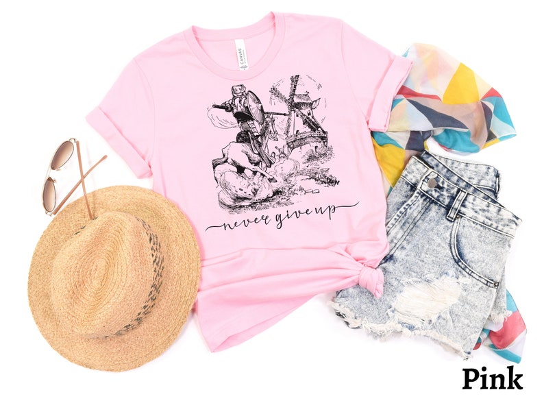 A pink shirt with the words never give up in a simple cursive font of black is lying on a white floor with shorts, straw hat, sunglasses & a scarf. Above that is an illustration of Don Quixote fighting a windmill.