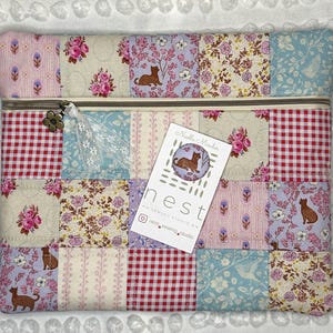 May include: A rectangular quilted zipper pouch featuring a patchwork design of floral, gingham, and cat-patterned fabrics in pastel pink, blue, and cream. A metal flower charm hangs from the zipper pull. A white product card labeled Needle Minder nest sewing studio is placed on the front.