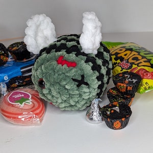 Puede incluir: Un juguete de peluche de crochet verde y negro con alas blancas, una boca roja cosida y una X negra cosida en la frente. El juguete está rodeado de varios dulces, incluyendo una bolsa de Fruit Patch Kids, un paquete de caramelos de chocolate y un caramelo de espiral rosa y blanco.