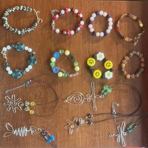 Puede incluir: Una colección de pulseras y collares de cuentas de colores, que incluyen una pulsera de cuentas blancas y negras, una pulsera de cuentas rojas y blancas, una pulsera de cuentas moradas, una pulsera de cuentas marrones, una pulsera de cuentas verdes y azules, una pulsera de cuentas amarillas, una pulsera de cuentas marrones y doradas, y un collar de alambre plateado con un dije de estrella.