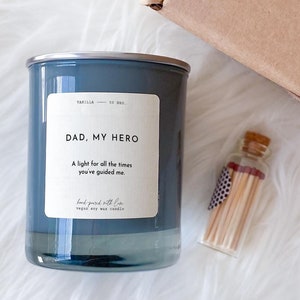 May include: A blue glass candle with a silver lid and the text "DAD, MY HERO" and "A light for all the times you've guided me."  The candle is next to a small glass jar with matches.