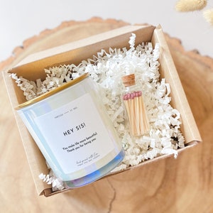 Op de afbeelding: Een cadeaudoos gevuld met wit geribbeld papier met daarin een heldere glazen kaars met een gouden deksel en een klein glazen potje met lucifers. De kaars heeft een wit etiket met de tekst "HEY SIS! You make life more beautiful. Thank you for being you." Het luciferspotje heeft een kurken deksel en roze luciferskopjes.