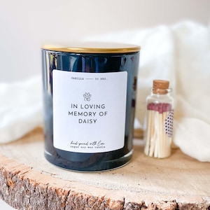 Puede incluir: Vela de cera de soja negra con tapa dorada y una etiqueta blanca que dice "Vanilla 50 hrs. In loving memory of Daisy hand poured with love vegan soy wax candle". Un pequeño frasco de vidrio con cerillas está al lado de la vela.