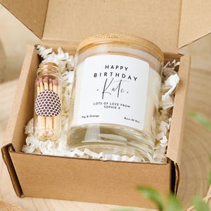 Puede incluir: Una caja de regalo con una vela de vidrio transparente con tapa de bambú. La vela tiene una etiqueta blanca con el texto "Happy Birthday Kate. Lots of love from Sophie X. Fig & Orange. Burn 35 Hrs." También hay un pequeño frasco de vidrio con cerillas en la caja.