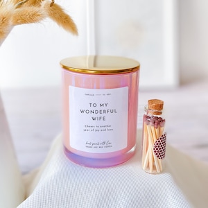 May include: Pink iridescent candle with a gold lid and a white label that reads "To My Wonderful Wife. Cheers to another year of joy and love. Hand poured with love. Vegan soy wax candle." A small glass jar with matches is next to the candle.