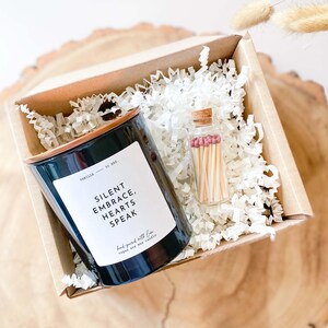 Peut inclure: Une bougie noire avec un couvercle en cuivre et une étiquette blanche qui dit "Silent Embrace, Hearts Speak" est dans une boîte brune avec du papier blanc déchiqueté. Un petit bocal en verre avec un couvercle en liège et des allumettes à l'intérieur est à côté de la bougie.