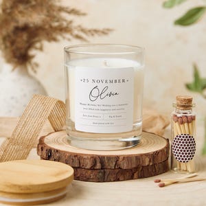 May include: A clear glass candle jar with a white label that reads "*25 NOVEMBER* Olivia Happy Birthday Sis! Wishing you a fantastic year filled with happiness and success. Love from [Name] Fig & Lotus hand poured with love" and a small glass jar with matches.
