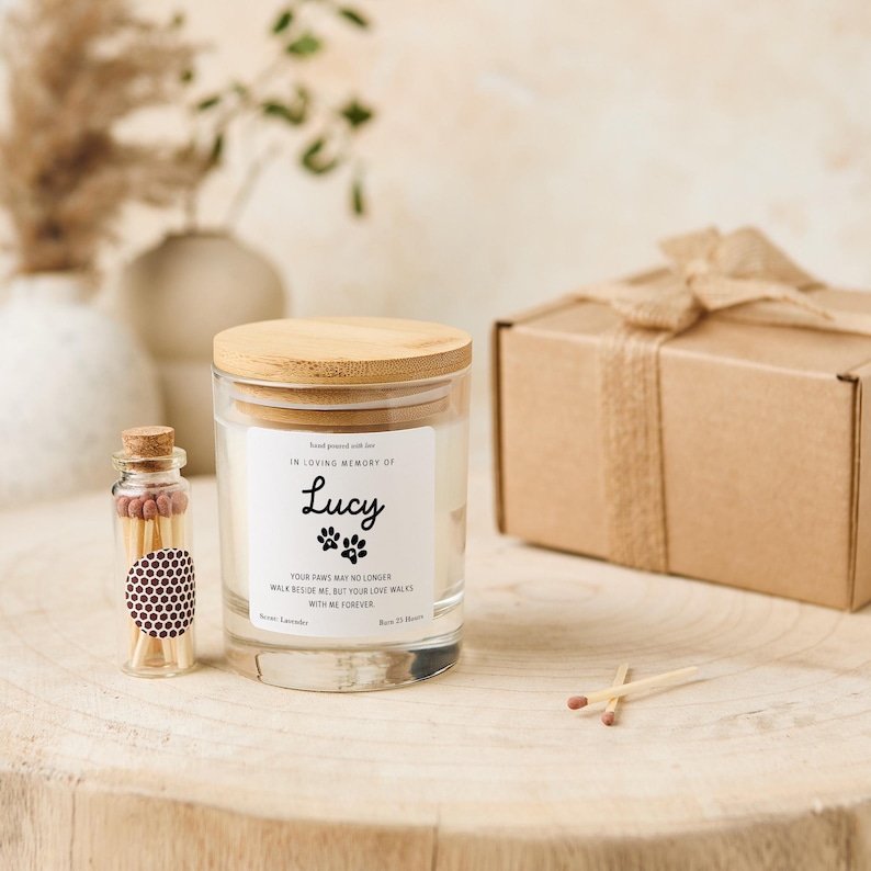 Pode incluir: Um frasco de vela de vidro transparente com tampa de madeira, um pequeno frasco de vidro com f&oacute;sforos e uma caixa de presente marrom amarrada com uma fita. A vela tem um r&oacute;tulo que diz "In Loving Memory of Lucy" com pegadas e uma mensagem.