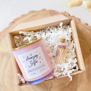 Op de afbeelding: Een geschenkdoos met een roze kaars met gouden rand en de tekst "Amazing Sister". Een klein glazen flesje met lucifers is ook inbegrepen. De kaars heeft een label met een boodschap en is omgeven door wit papier.