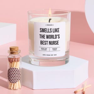 Könnte beinhalten: Eine brennende Duftkerze in einem durchsichtigen Glas mit dem Text "SMELLS LIKE THE WORLD'S BEST NURSE" auf einem weißen Etikett. Die Kerze steht auf einer weißen Plattform, links daneben eine kleine Glasflasche mit Streichhölzern. Hergestellt aus 100 % veganem Sojawachs.