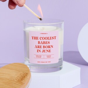 May include: A lit match ignites a scented candle in a clear glass. The candle has a pink label with the text "THE COOLEST BABES ARE BORN IN JUNE." The candle is made of 100% vegan soy wax and has a burn time of 25 hours.