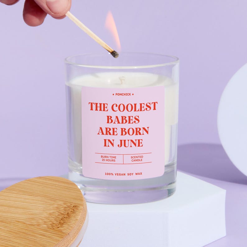 May include: A lit match ignites a scented candle in a clear glass. The candle has a pink label with the text "THE COOLEST BABES ARE BORN IN JUNE." The candle is made of 100% vegan soy wax and has a burn time of 25 hours.