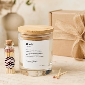 Puede incluir: Un tarro de vela de vidrio transparente con tapa de madera y una etiqueta que dice "Bestie". También se ven una pequeña botella de vidrio de cerillas y una caja de regalo. La vela tiene aroma a lavanda.