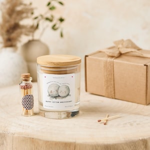 May include: A glass candle jar with a wooden lid, a small glass bottle of matches, and a wrapped gift box. The candle has a label that reads "Happy Cotton Anniversary" with a cute illustration. The candle is scented with Seven Lavender and has a 25-hour burn time.
