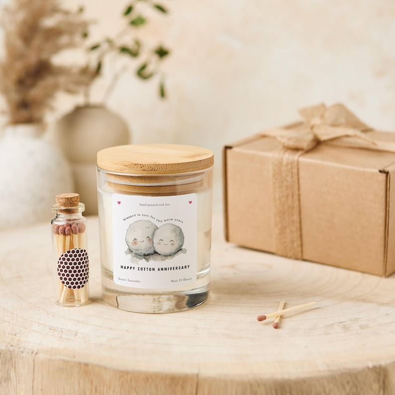 May include: A glass candle jar with a wooden lid, a small glass bottle of matches, and a wrapped gift box. The candle has a label that reads "Happy Cotton Anniversary" with a cute illustration. The candle is scented with Seven Lavender and has a 25-hour burn time.