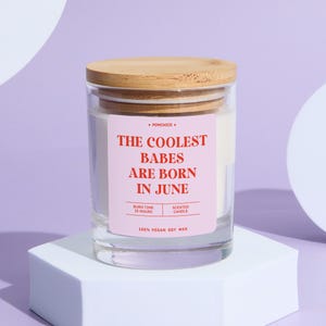 May include: A clear glass candle jar with a wooden lid. The jar has a pink label with the text "THE COOLEST BABES ARE BORN IN JUNE". The candle is on a white geometric stand against a purple background.
