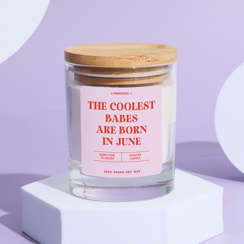 May include: A clear glass candle jar with a wooden lid. The jar has a pink label with the text "THE COOLEST BABES ARE BORN IN JUNE". The candle is on a white geometric stand against a purple background.